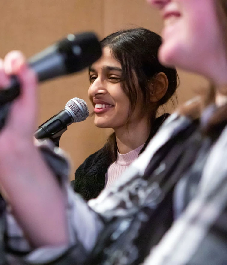 A student singer in a pink sweater smiling while singing into a microphone during a band practice session. A student singer in a pink sweater smiling while singing into a microphone during a band practice session.